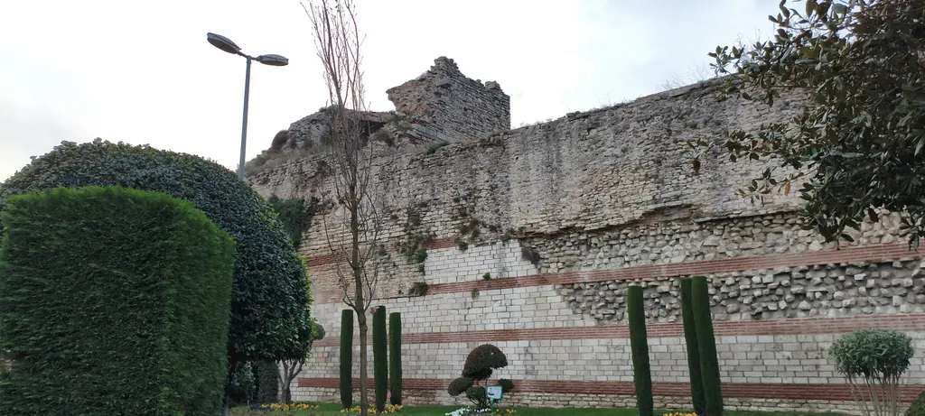 Uzun bir sur bölümü; taş ile tuğlanın almaşık sıraları, yıkık üst kenar, önünde budanmış çalılar ve bakımlı yeşil şerit.