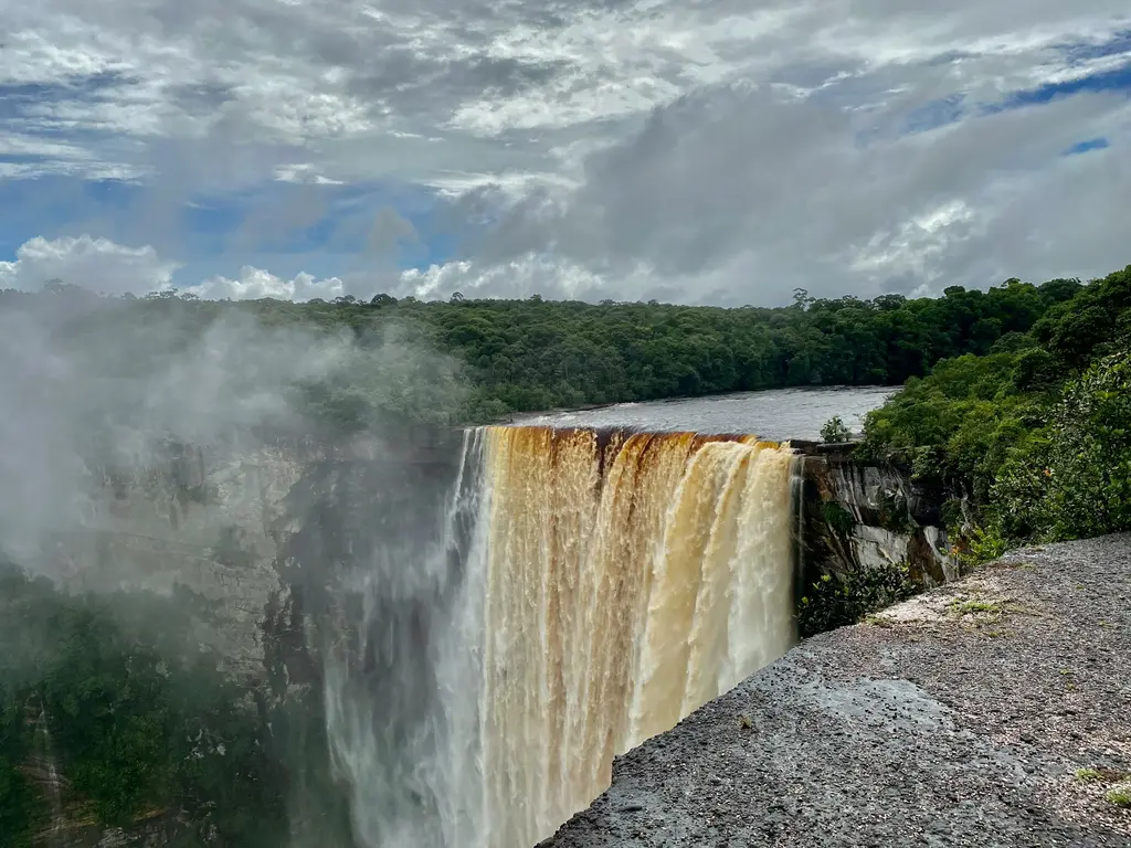 Guyana'da Kaieteur Şelalesi'nin uçurumdan tek parça hâlinde dökülen tanen renkli kahverengi suları; yükselen su buharı, çevredeki yoğun yağmur ormanı ve bulutlu gökyüzü.