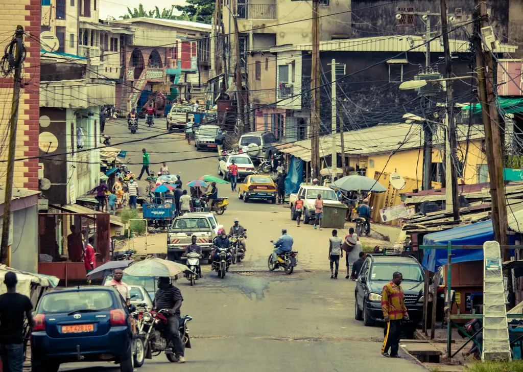 Duala'da eğimli, kalabalık bir cadde; arabalar, motosikletler, pazar şemsiyeleri, yayalar ve havada birbirine karışmış elektrik telleri.