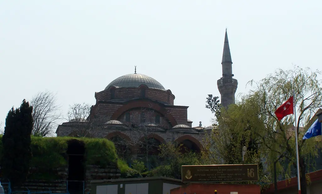 Rum Mehmed Paşa Camii; bahçe ve servi ağaçlarının ardında kubbe, tuğla kemerler, minare ve bayrak direği.