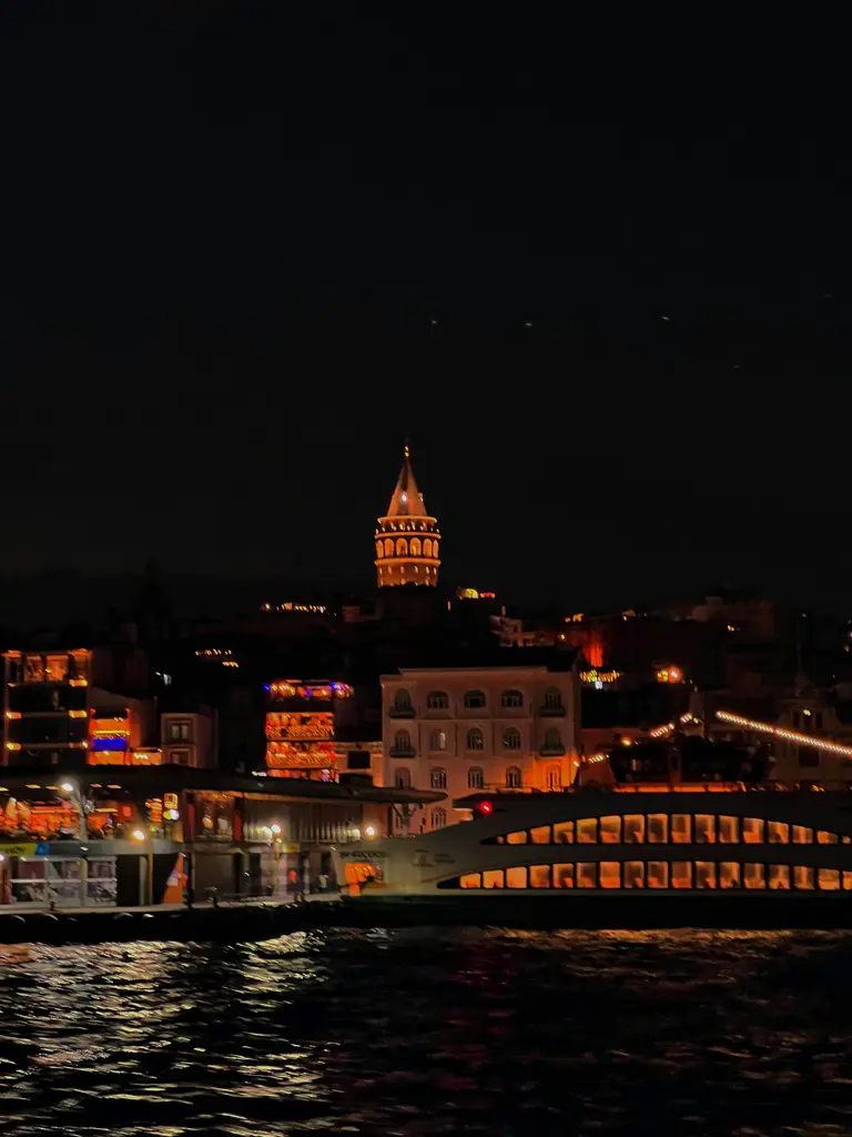 Gece denizden Galata; yamaçtaki ışıklı binalar, tepede aydınlatılmış Galata Kulesi ve suya düşen yansımalar.