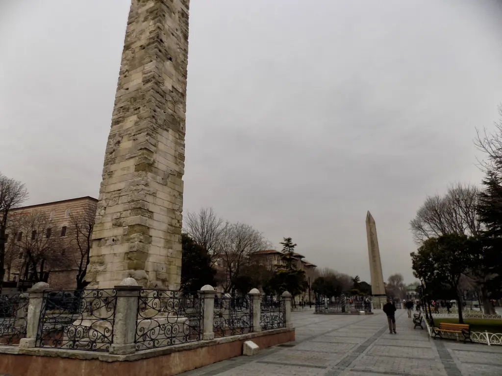 Önde Örme Sütun'un taş gövdesi, arkada uzakta Dikilitaş; parmaklık, yapraksız ağaçlar ve bulutlu kurşuni gökyüzü.