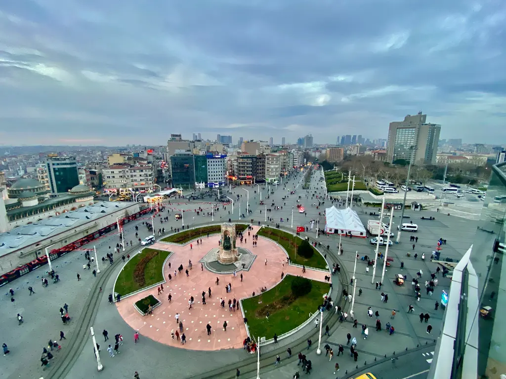 Yukarıdan Taksim Meydanı; ortada yeşil çimlerle çevrili pembe taş döşeli alan ve anıt, çevresinde yürüyen kalabalık, arkada Beyoğlu'nun binaları.