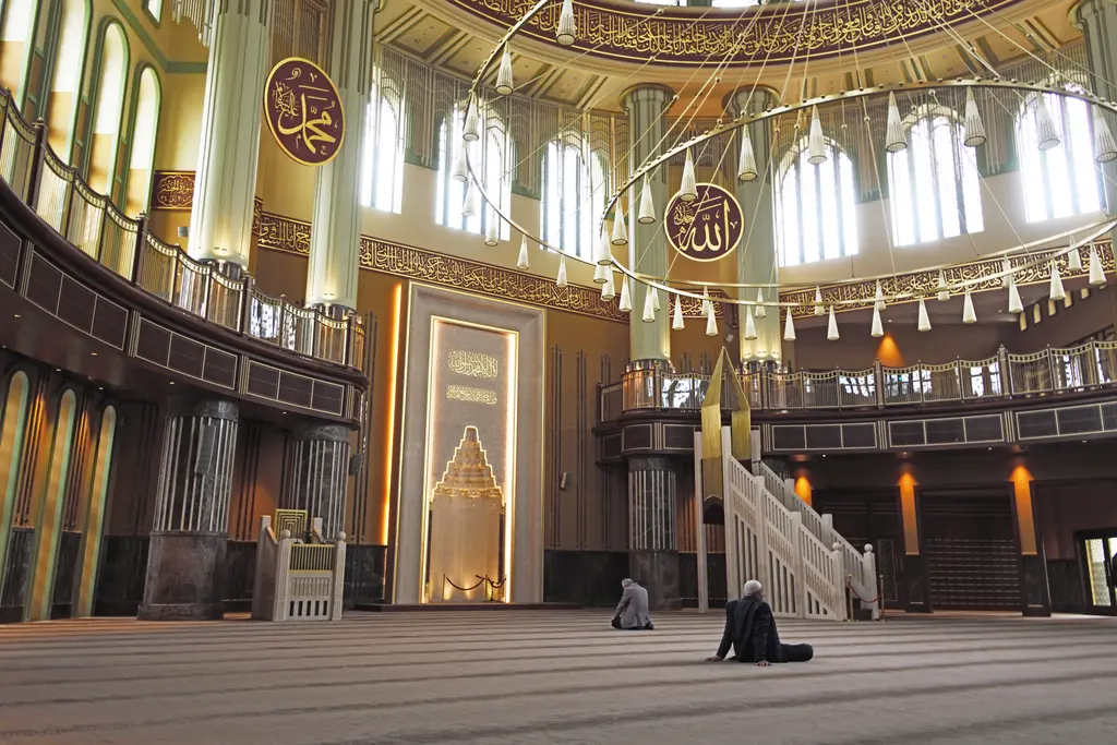 Taksim Camii'nin harimi; altın tonlu kubbe, galerilerdeki kemerli pencere dizisi, asılı avize çemberi ve ortada beyaz mihrap.