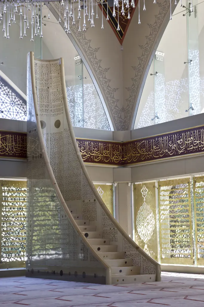 Şakirin Camii'nin minberi; beyaz, şeffaf akrilik basamaklar, kıvrılarak yükselen gövde ve arkada cam paneller.