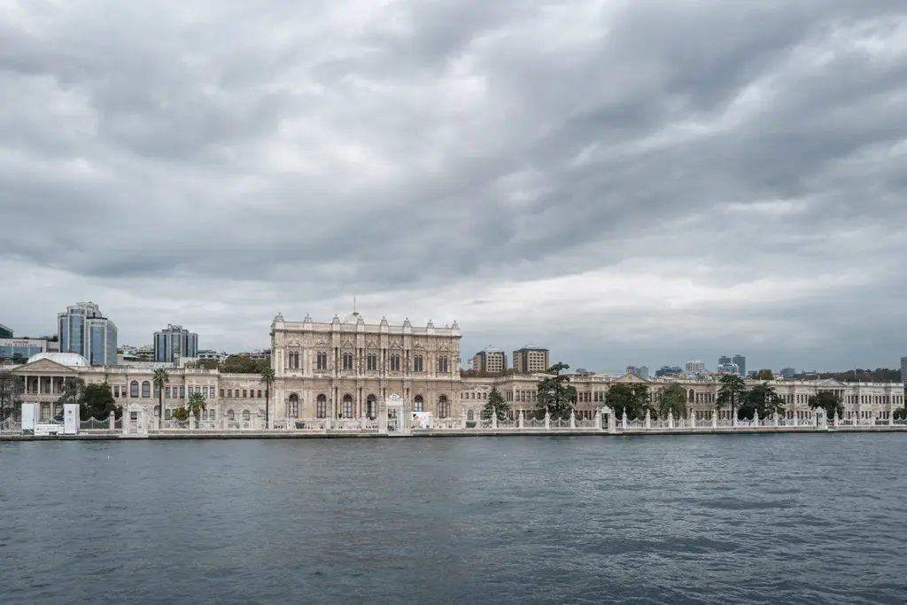 Boğaz'dan Dolmabahçe Sarayı; bulutlu gökyüzü altında uzun beyaz saray cephesi, kıyıdaki rıhtım ve arkada yamaca yayılan binalar.