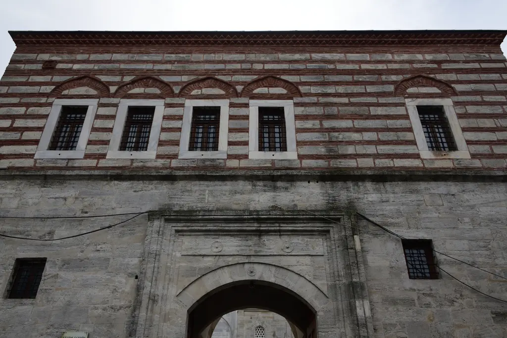 Yeni Valide Külliyesi'nde mektep; taş ve tuğla sıralı cephe, sıra pencereler ve altta kemerli geçit.