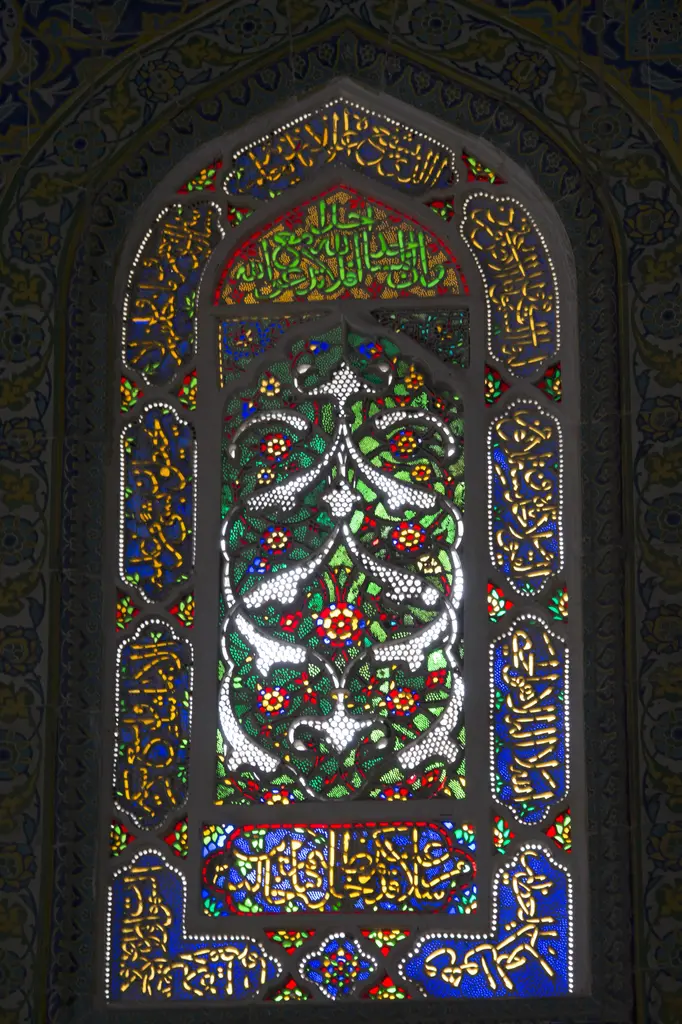 Şehzade Mehmed Türbesi'nde renkli cam pencere; beyaz, kırmızı ve yeşil camlardan kurulmuş çiçekli ağaç motifi, üstte ve altta hat panoları, mavi çini çerçeve.
