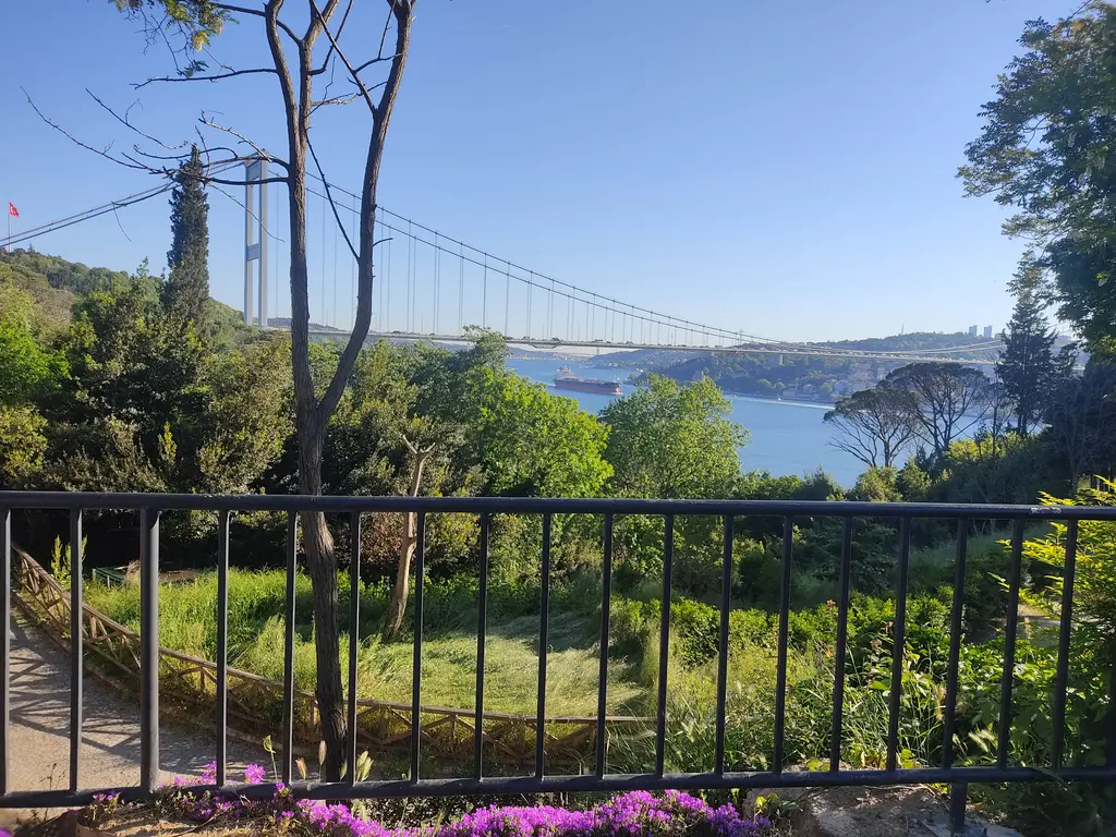 Mihrabat Tabiat Parkı'nın girişinde demir korkuluklu seyir yeri; önde mor çiçekler, ortada ağaçlar, arkada Boğaz ve köprü.
