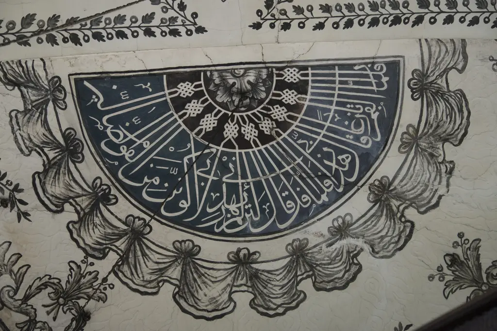Duvarda kalem işi pano; yarım daire biçiminde, çizgilerle bölünmüş yazılı alan ve çevresinde siyah beyaz çelenk ile yaprak motifleri.