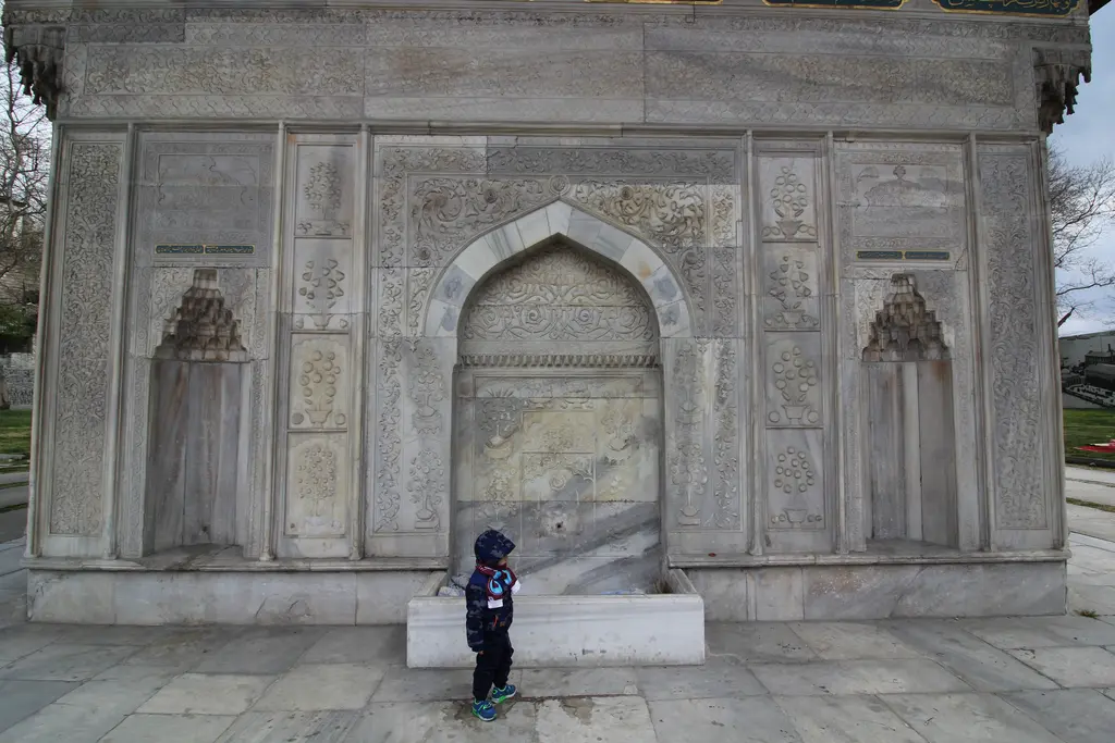 Tophane Çeşmesi'nin bir cephesi; mermer üzerinde kabartmalı nişler, ortada kemerli musluk ve önünde bir çocuk.