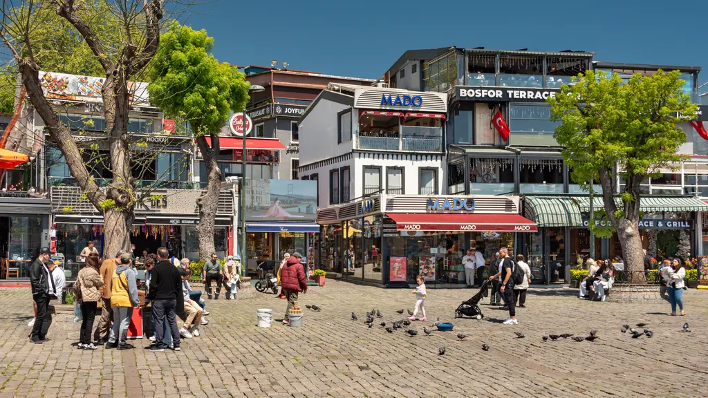Ortaköy meydanında yürüyen insanlar ve güvercinler; taş döşeme, ağaçlar, cumbalı renkli evler ve kafe tabelaları.