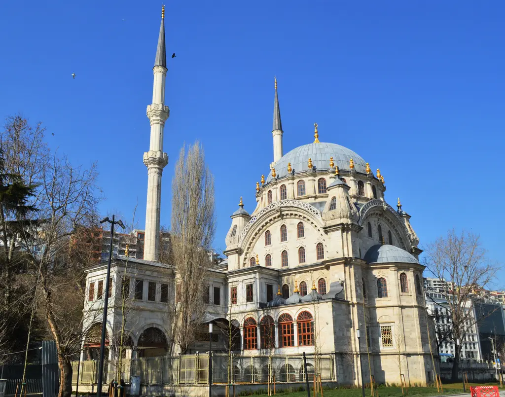 Tophane'de Nusretiye Camii; kurşun kaplı kubbe, çevresinde altın yaldızlı alemler, iki ince minare, önde iki kanatlı hünkâr kasrı ve ağaçlar.