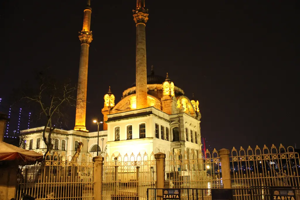 Gece Ortaköy Camii; altın sarısı ışıkla aydınlatılmış cephe ve minareler, önde demir parmaklık ve karanlık gökyüzü.