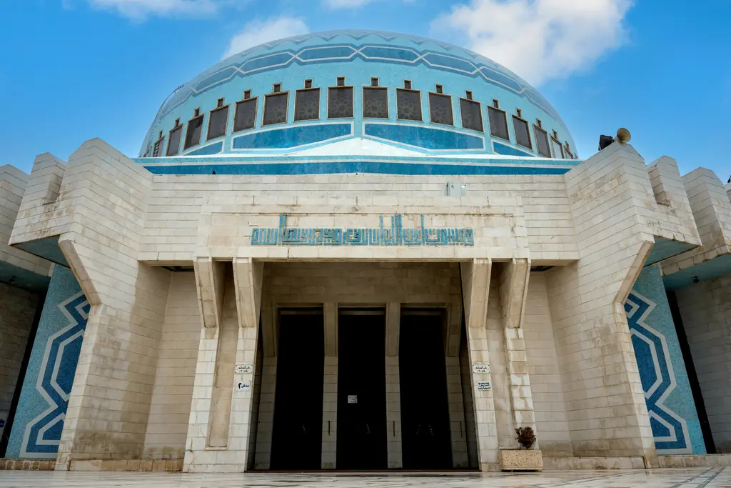 Amman'da caminin cephesi; mavi mozaik karolarla kaplı büyük kubbe, açık renkli taş duvarlar, geometrik desenli mavi süslemeler ve ortadaki anıtsal giriş.