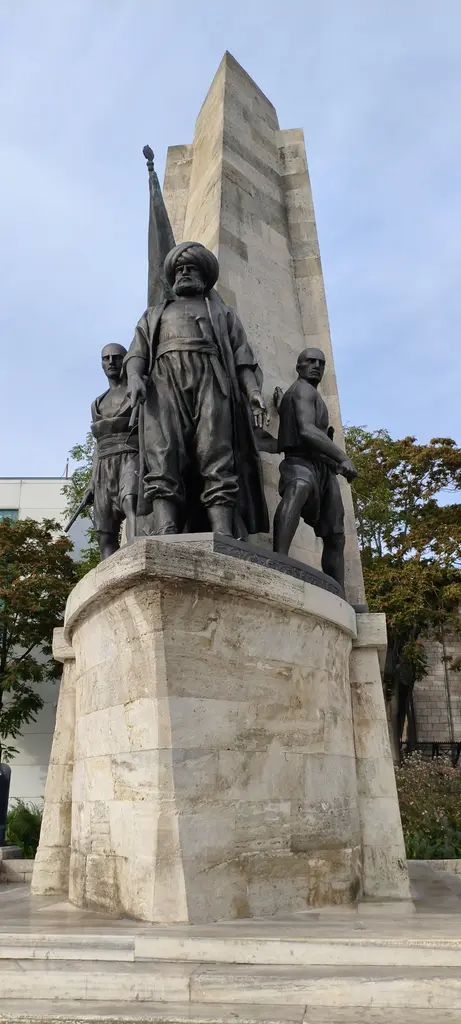 Barbaros Anıtı; önden bakışta taş kaide üzerinde üç bronz figür ve arkalarında sivri uçlu yüksek taş kütle.