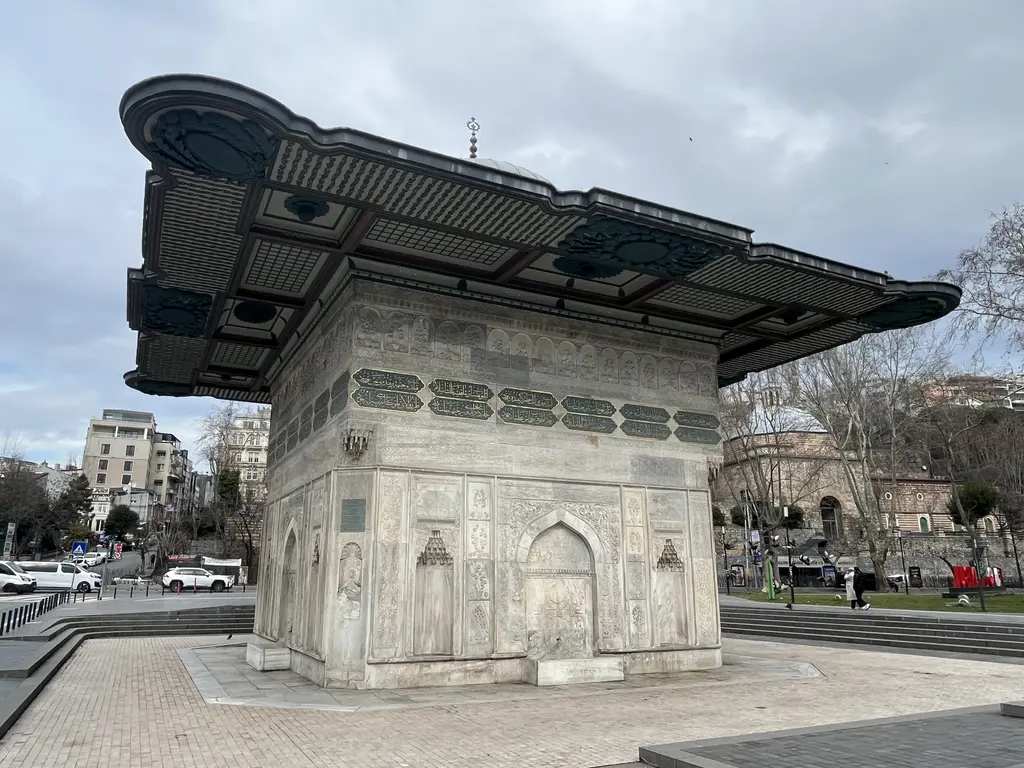Tophane Çeşmesi; geniş saçaklı kare mermer gövde, kabartmalı cepheler ve çevresindeki taş döşeli meydan.