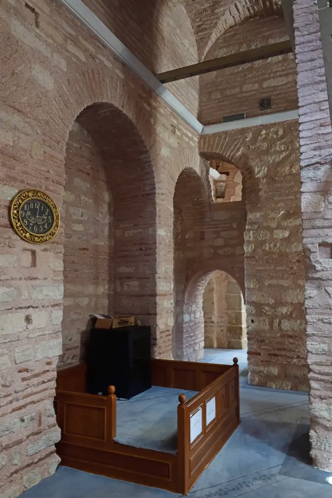 Fenari İsa Camii'nde dehliz; sıvası sökülmüş tuğla ve taş payeler arasında kemerli geçitler, solda yaldız çerçeveli duvar saati ve ahşap korkuluklu alçak platform.