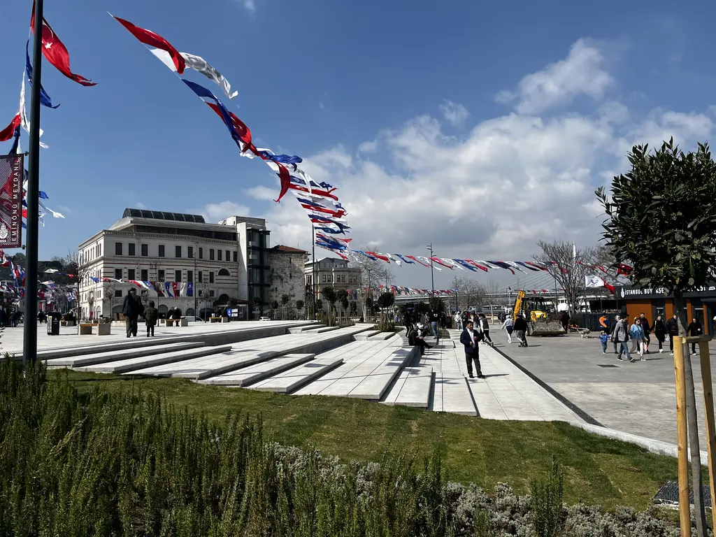 Eminönü meydanında yeşil alan ve taş basamaklar; yürüyen insanlar, flamalar, ağaçlar ve arkada taş bina.