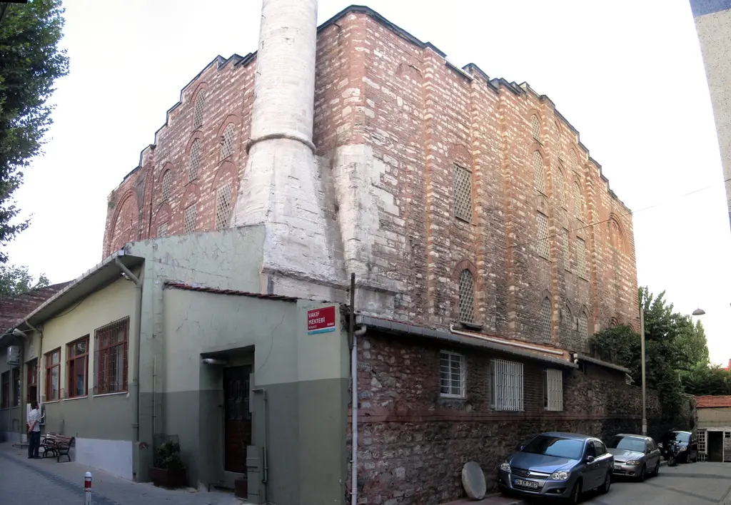 Gül Camii'ne sokak köşesinden bakış; alçak müştemilatın üstünde yükselen tuğla gövde ve minare, park etmiş arabalar, bankta oturan bir kişi.