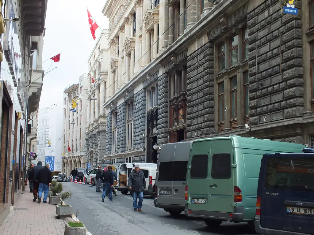 Bankalar Caddesi; sağda kesme taş cepheli büyük bina, kaldırımda yürüyen insanlar, yolda park etmiş minibüsler ve bayraklar.