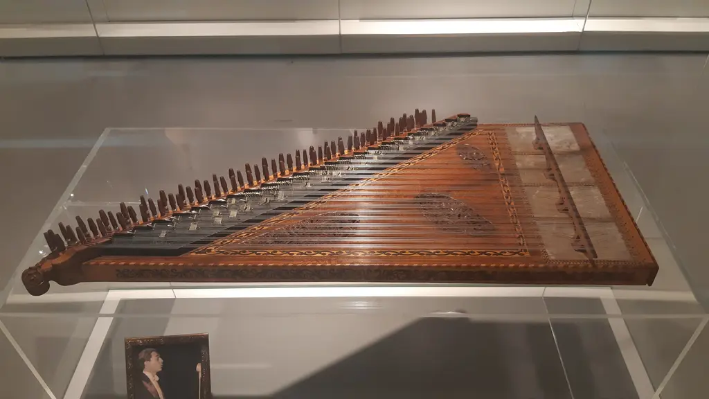 AKM Müzik Platformu'nda cam vitrinde bir kanun; ahşap gövde, uzun tel dizisi, akort mandalları ve altta bir portre fotoğrafı.