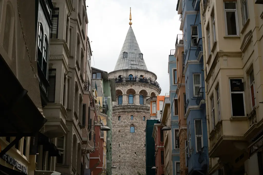 Beyoğlu'nda iki apartman arasından Galata Kulesi; cumbalı eski cepheler, dar sokağın gölgesi ve arkada beyaz gökyüzü.