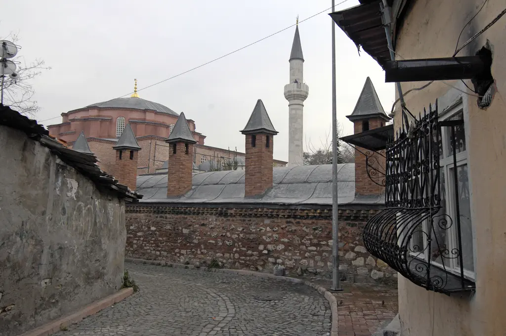 Küçük Ayasofya'da sisli bir sokak; solda taş tuğla avlu duvarı ve üzerinde konik taş külahlar, arkada caminin kubbesi ile minaresi, sağda beyaz badanalı ev cephesi.