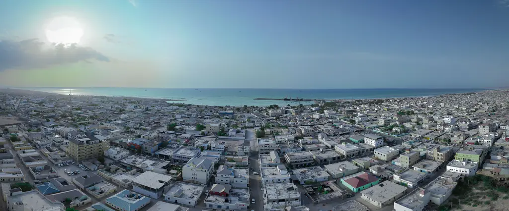 Bosaso'da havadan panoramik görünüm; ufka kadar uzanan beyaz ve bej düz damlı binalar, arkada Aden Körfezi ve pusla yumuşamış ufuk, solda parlayan güneş.