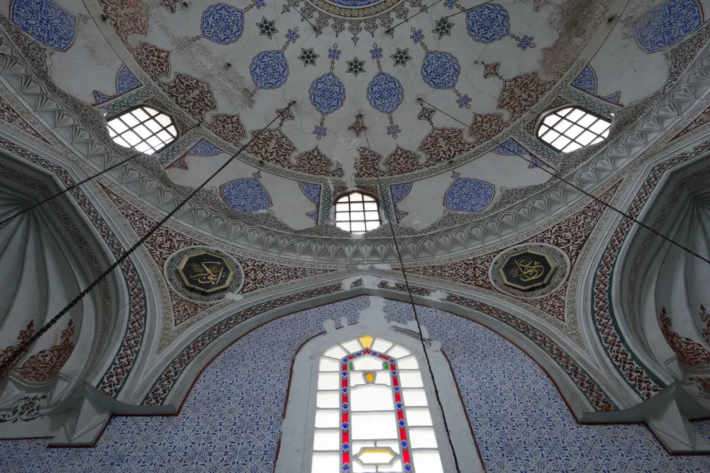 Haseki Sultan Camii'nde kubbe eteği; mavi kalem işi süslemeler, madalyonlar, yuvarlak pencereler ve altta renkli camlı bir pencere.