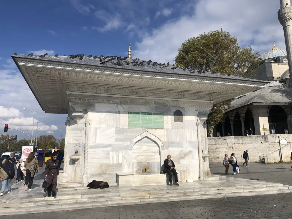 Üsküdar III. Ahmed Çeşmesi; çatısına konmuş güvercinler, mermer cephe, yanında oturan insanlar ve cami revakı.