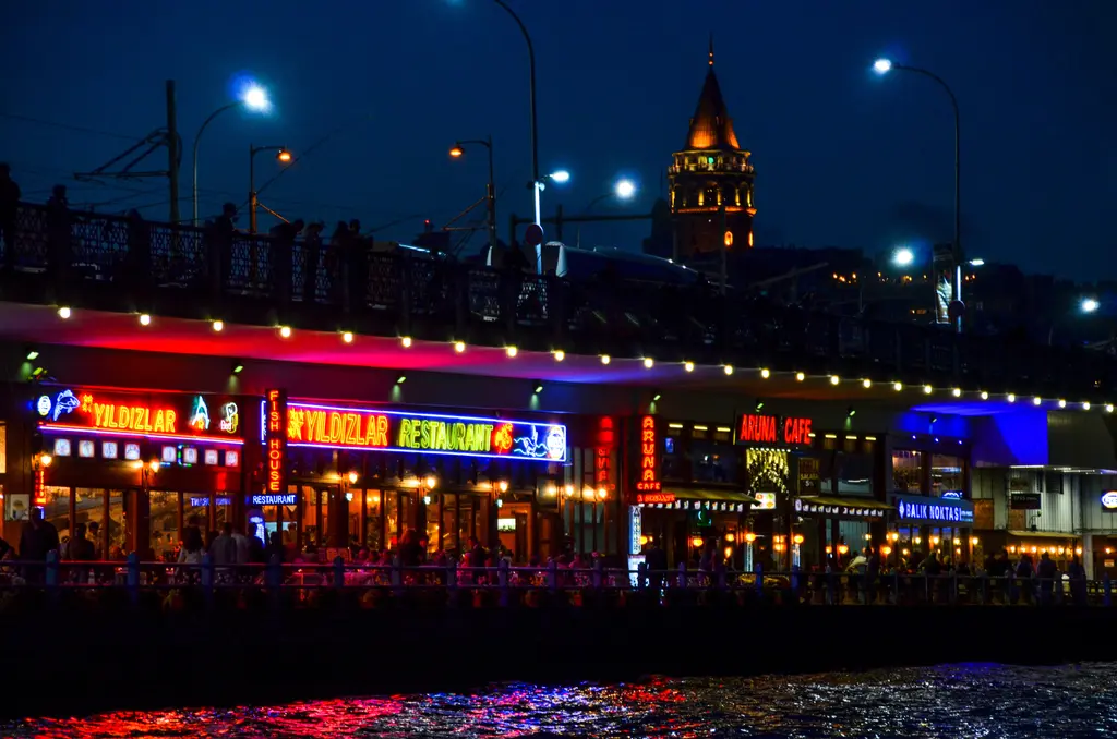 Gece Galata Köprüsü'nün alt katı; renkli ışıklı lokanta tabelaları, suya yansıyan ışıklar ve arkada Galata Kulesi.