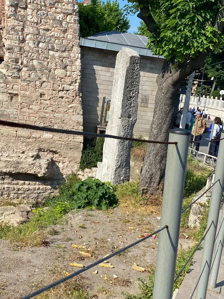 Milyon Taşı; solda moloz taş örgülü duvar, ortada mermer ayak, önde metal korkuluk ve sağda kaldırımda yürüyen ziyaretçiler.