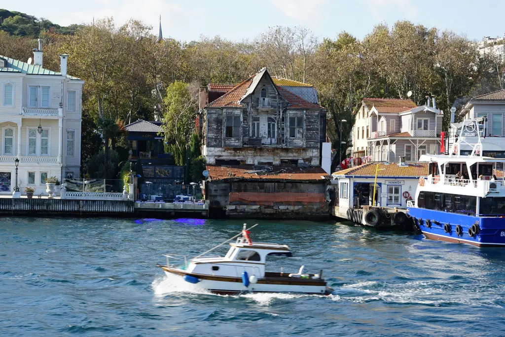 Yeniköy İskelesi çevresi; eski ahşap yalı, iskeleye yanaşmış mavi tekne, önde geçen beyaz motor ve arkada ağaçlar.