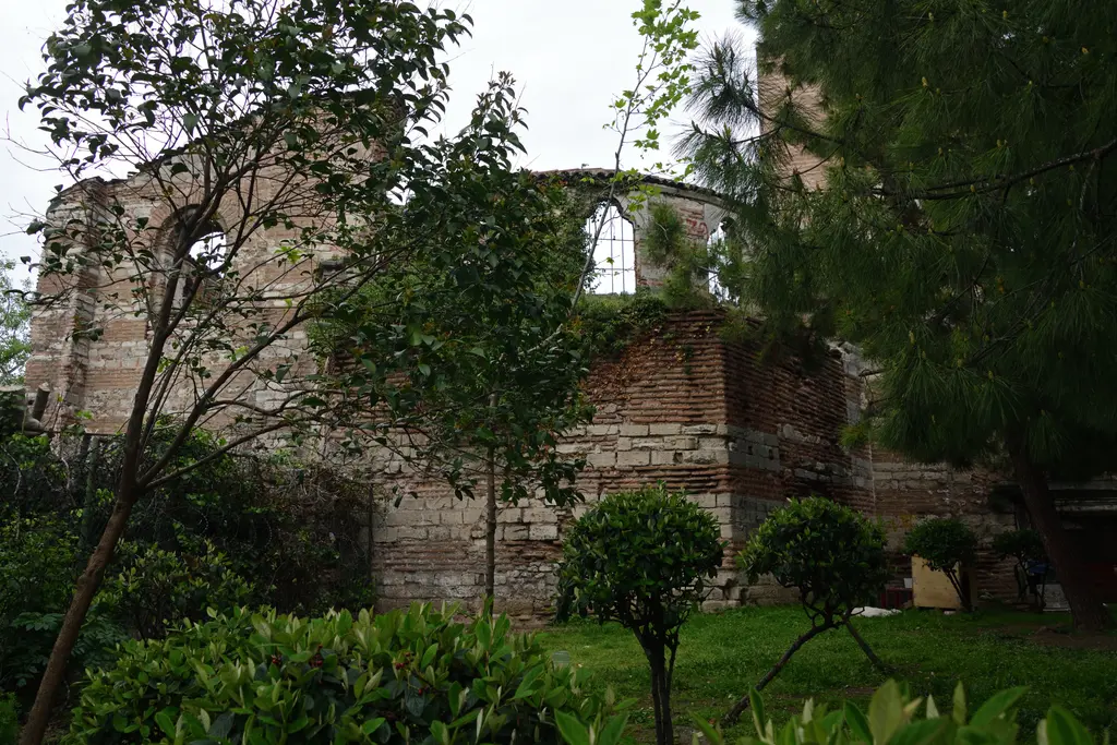İmrahor İlyas Bey Camii'nin bahçesi; budanmış çalılar ve genç ağaçların arkasında kemerli pencereli yıkık duvarlar, sağda kiremitli saçak, gri bulutlu gökyüzü.