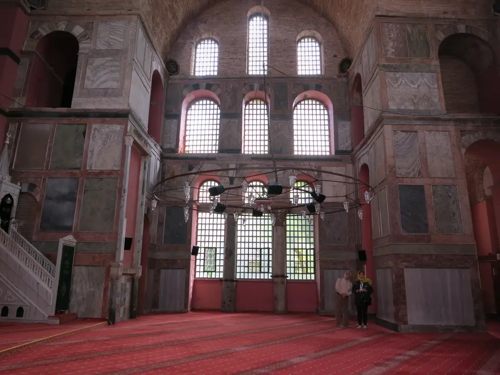 Kalenderhane Camii'nin harimi; kemerli pencerelerle dolu yüksek duvar, renkli damarlı mermer kaplı ayaklar, halka avize ve iki ziyaretçi.