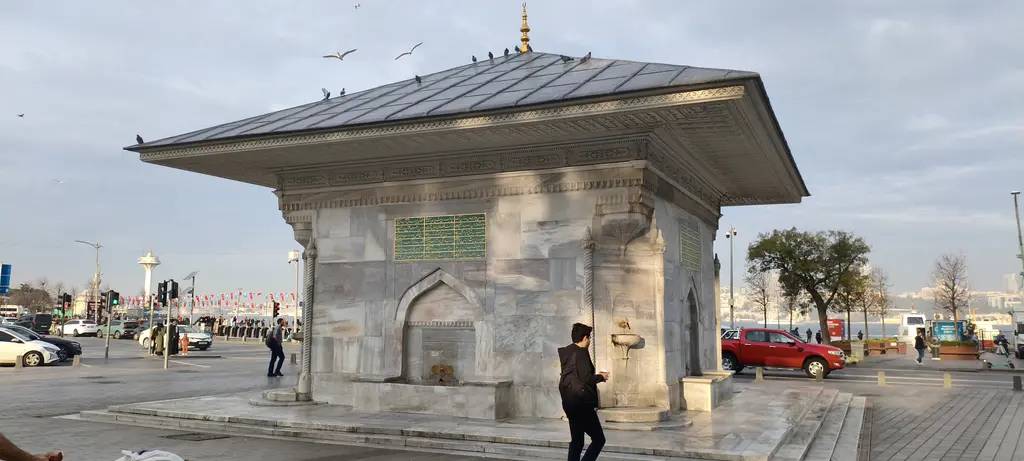 Üsküdar III. Ahmed Çeşmesi; geniş meydanda kare gövdeli çeşme, çatıda kuşlar, kıyıda iskele ve Boğaz.