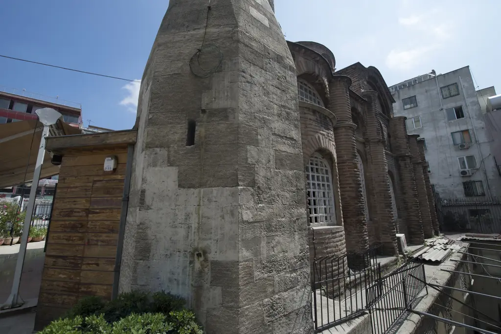 Bodrum Camii'nde önde minarenin masif kesme taş kaidesi, arkasında güney cephesi; sıralı payandalar, kafesli kemerli pencereler, sağda alt yapıya inen merdiven.