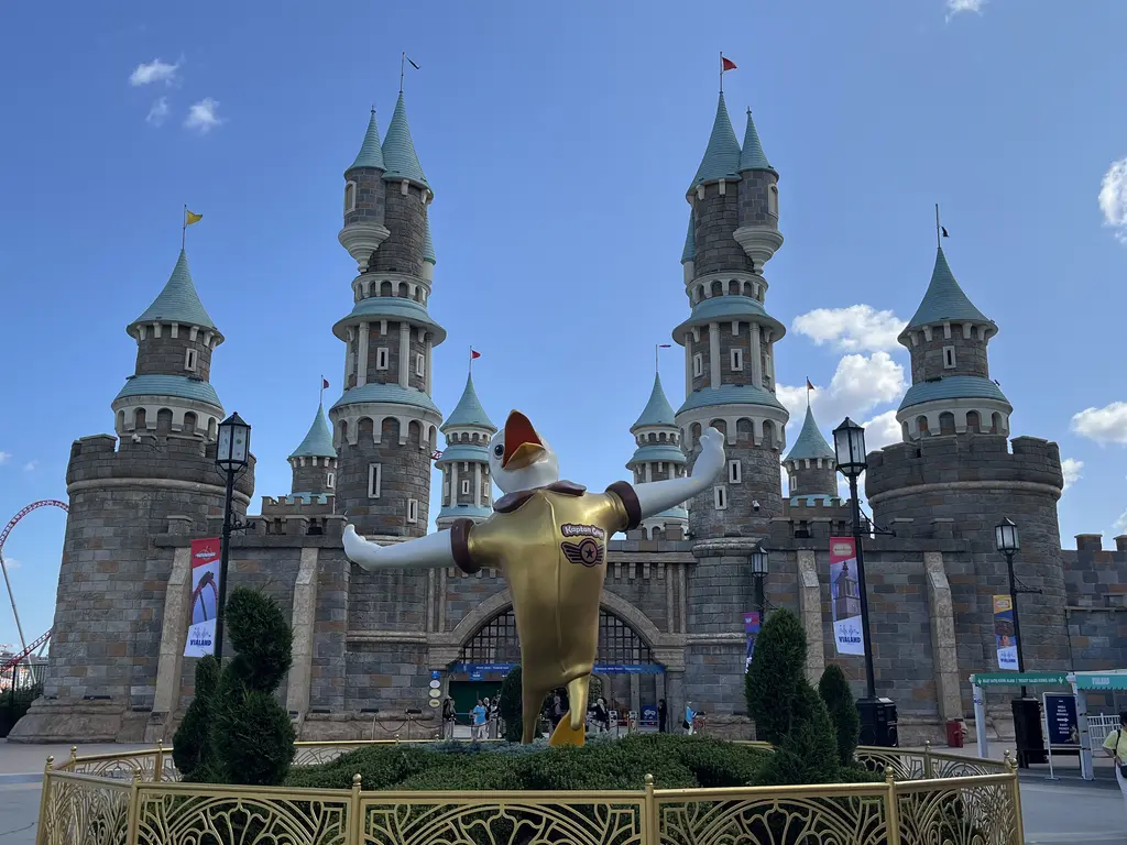 Vialand'de gri taş kuleli, turkuaz külahlı masalsı şato; önünde kollarını açmış altın renkli maskot heykeli, çiçekli çit ve yürüyen ziyaretçiler.