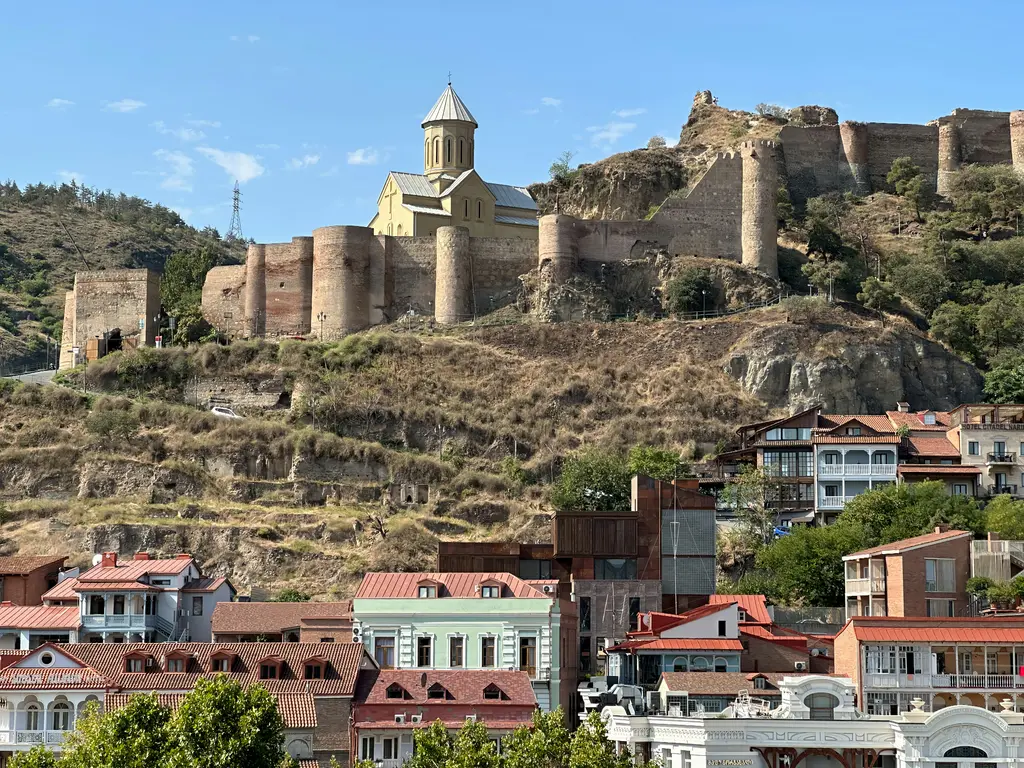 Tiflis'te Narikala Kalesi'nin taş surları ve kuleleri; arkasında şehir ve yeşil yamaçlar.
