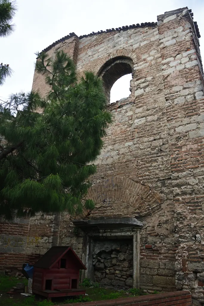 İmrahor İlyas Bey Camii'nin doğu cephesi; altta mermer sövesi duran örülmüş kapı, üstünde tuğla hafifletme kemeri, yukarıda apsis kasnağı, solda çam ağacı.