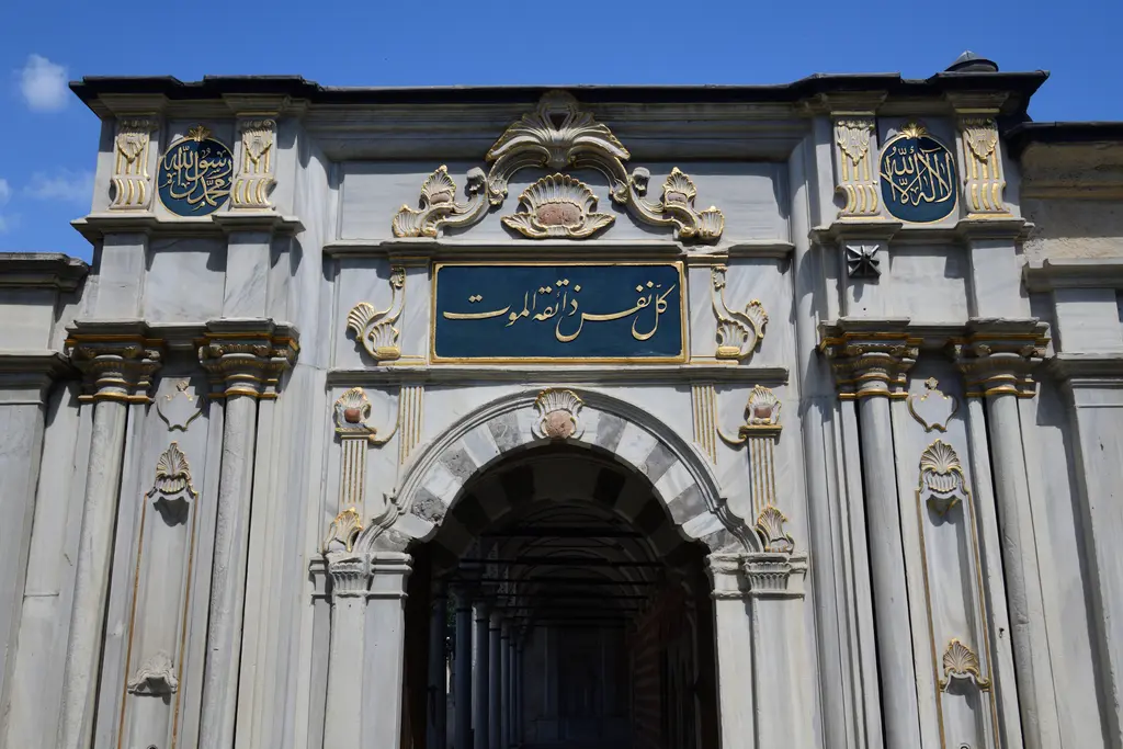 Mihrişah Valide Sultan Külliyesi'nin hazire kapısı; iki renk mermer kemer, mavi zeminli yazı panosu, yaldızlı madalyonlar ve mavi gökyüzü.