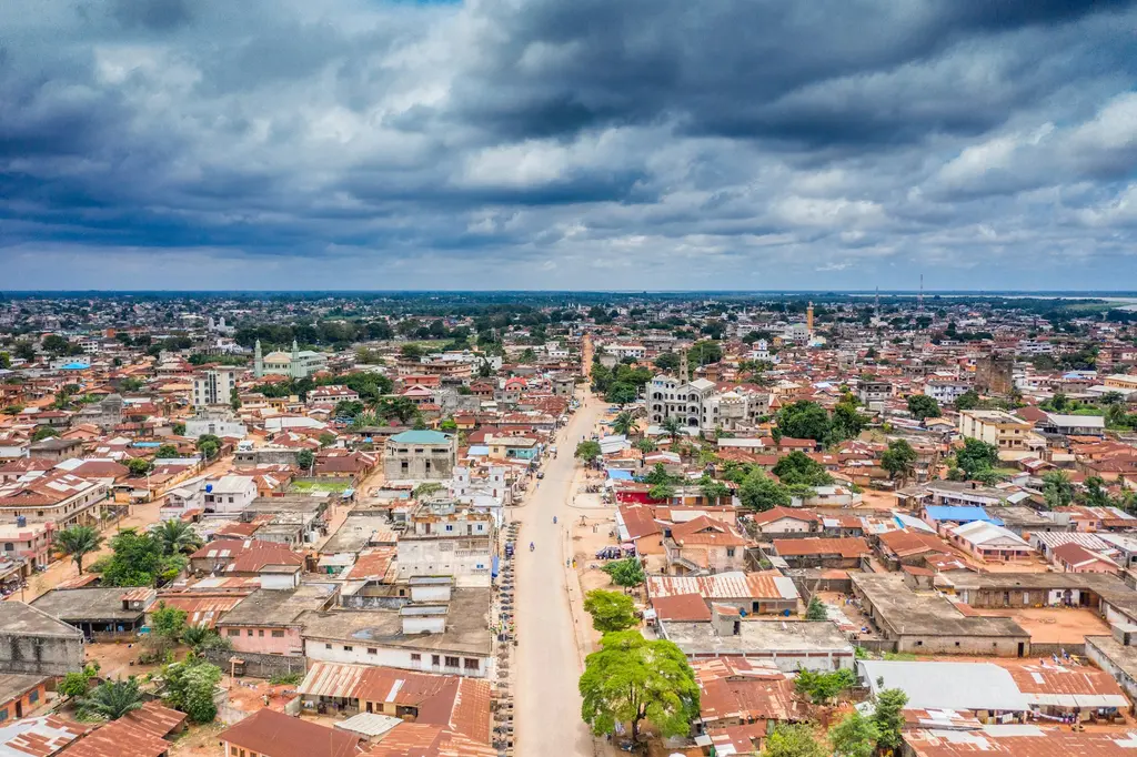 Benin'de kızıl teneke çatılı evlerin ufka dek uzandığı bir kentin havadan görünümü; ortadan geçen geniş bir cadde, minareli camiler ve yağmur bulutlu gökyüzü.