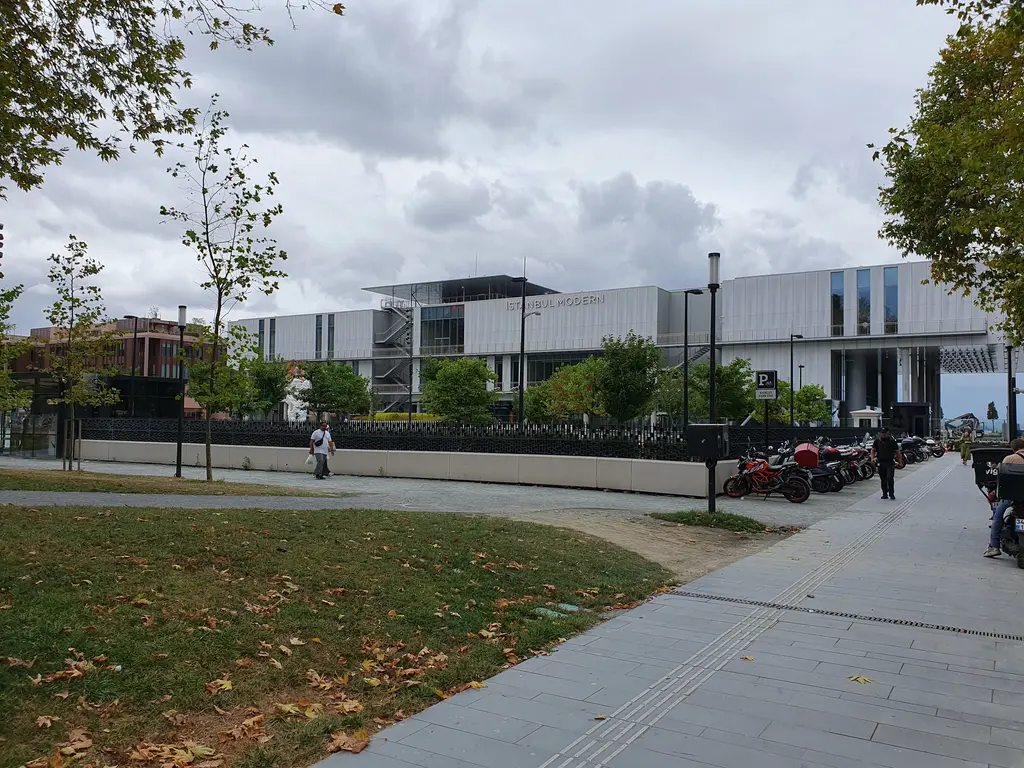 Çimenli bir parkın kenarından İstanbul Modern; önde ağaçlar ve yürüyüş yolu, arkada alçak gri bina.