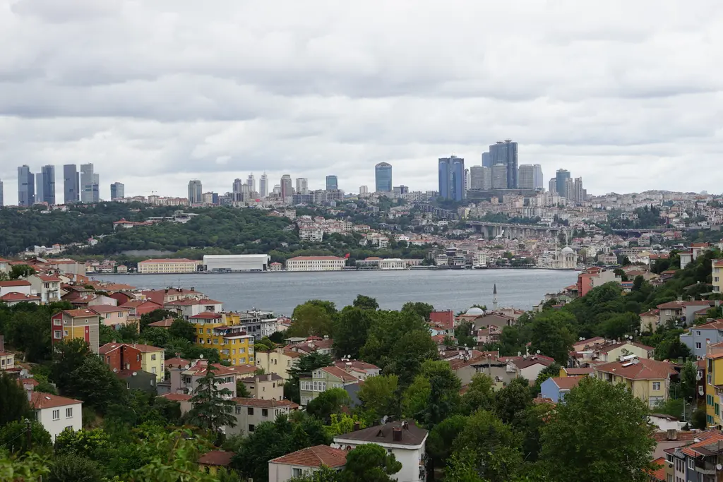 Kuzguncuk'tan Boğaz; önde ağaçlar arasında kiremit çatılı evler, ortada mavi su ve karşı kıyıda yüksek binalar.