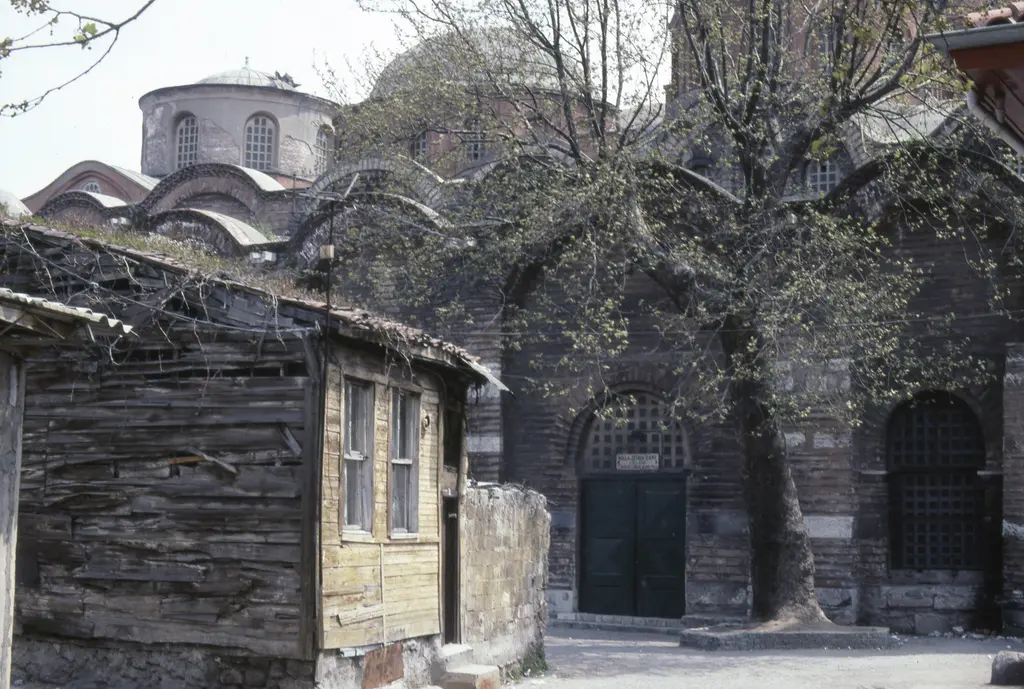 Zeyrek Camii ile yanındaki gri ahşap ev; solda cumbalı eski konut, sağda demir kapı ve kubbeli tuğla gövde, önde yapraksız ağaç dalları.