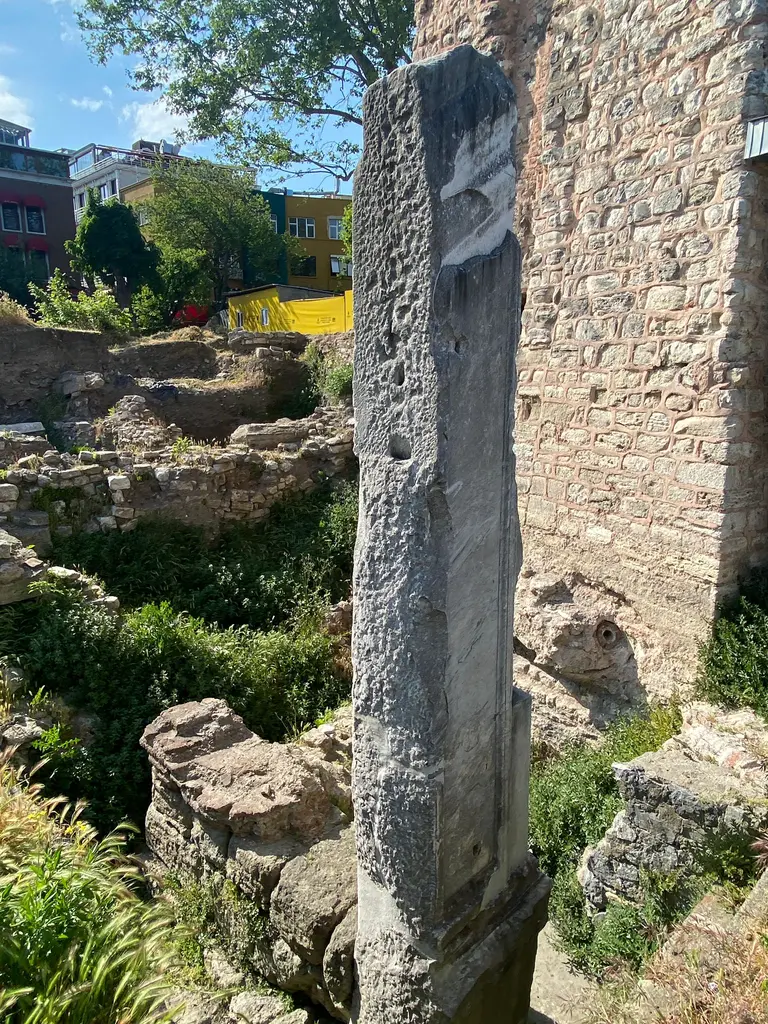Milyon Taşı'nın mermer ayağı; arkasında tuğla örgülü duvar, solda otlar arasında kazıdan çıkan taş kalıntılar ve eski binalar.