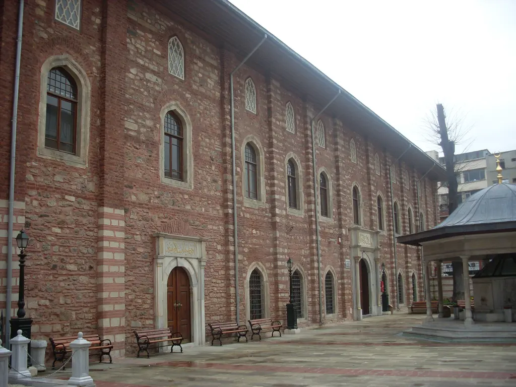 Arap Camii'nin avlusu; uzun tuğla ve taş cephe, iki sıra pencere, sağda kubbeli şadırvan, önde taş döşeli geniş avlu.