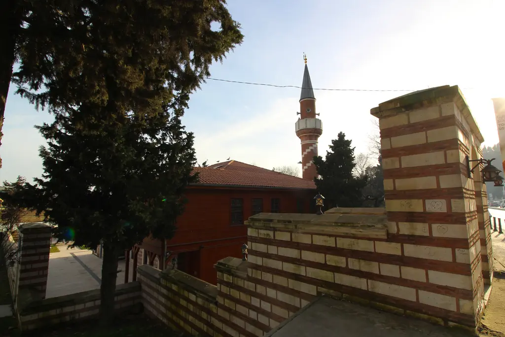 Şah Sultan Camii'nin avlusu; tuğla-taş hazire duvarı, çamlar, kiremit çatı ve arka planda tuğla minare.