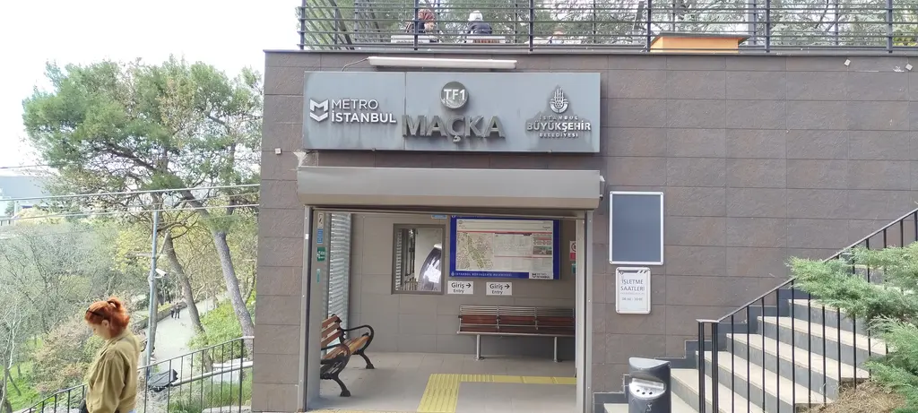 Maçka Demokrasi Parkı içinde TF1 Maçka istasyonunun girişi; gri taş cephede Metro İstanbul tabelası, banklı bekleme yeri ve merdiven.